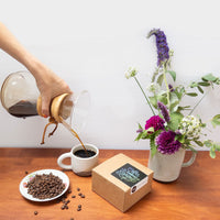 A hand holding a chemex brewer pouring coffee into a mug, on a table with the Swelter Coffee Gift Box next to it, and a vase with flowers