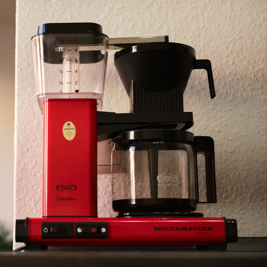 A red Moccamaster coffee brewer on a table in front of a white wall